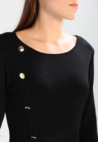 Pull noir en tricot côtelé avec un large décolleté en U et des accents de boutons dorés sur le côté. Texture lisse et coupe ajustée.