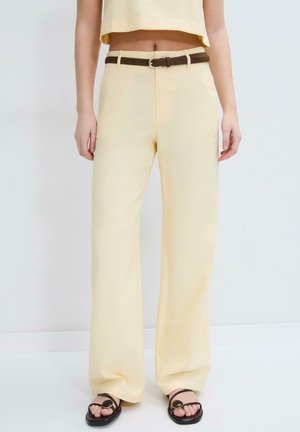 Ženska, oblečena v kremne široke hlače s rjavim pasom in črnimi sandali z okroglimi zlatimi poudarki na belem ozadju.