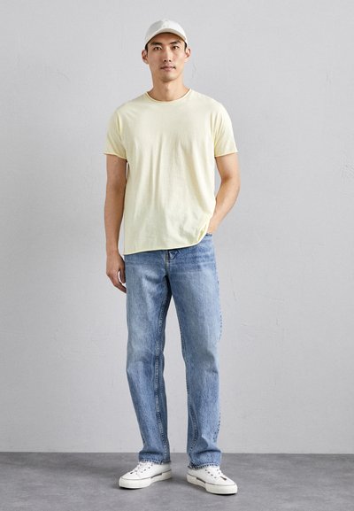 T-shirt en coton jaune clair, jeans en denim bleu et baskets blanches. La tenue est complétée par une casquette de couleur claire. Style simple et décontracté.