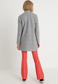 Šedý vlněný kabát s rovným střihem, kombinovaný s červenými pruhovanými rozšířenými kalhotami. Výrazné textury zahrnují hladký povrch kabátu a vzor látky kalhot.