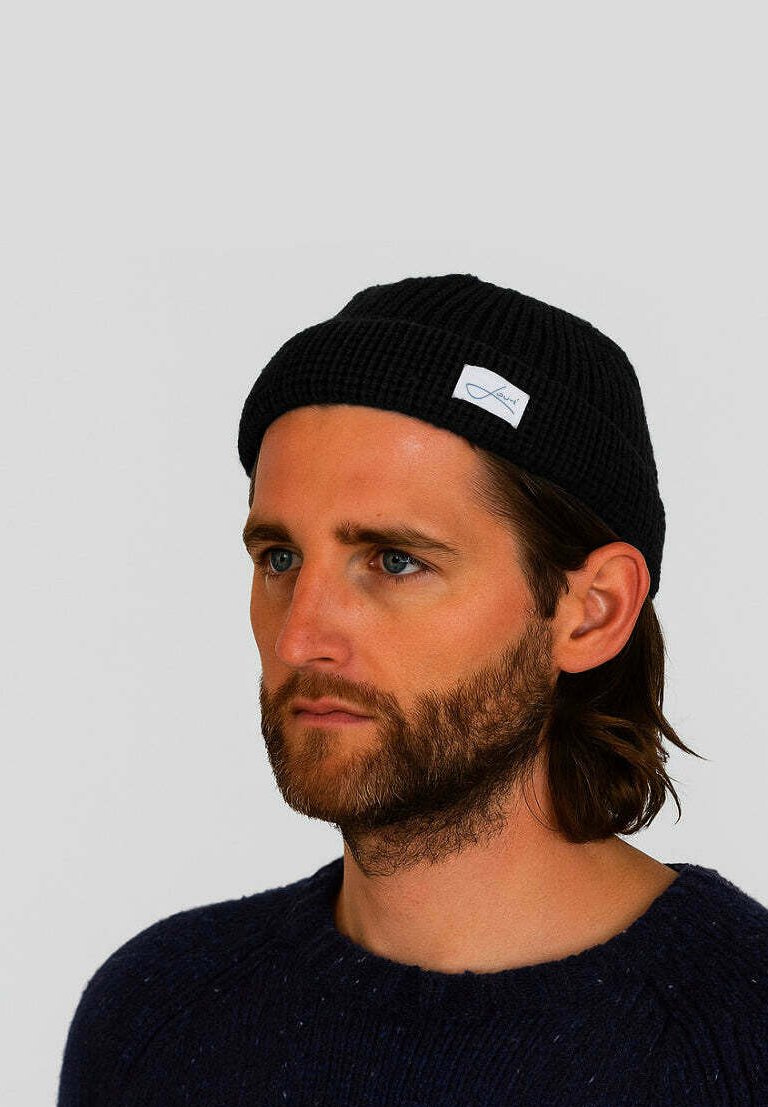Black knitted beanie with a ribbed texture, featuring a small white label. Person wearing it has a neutral expression and long hair.