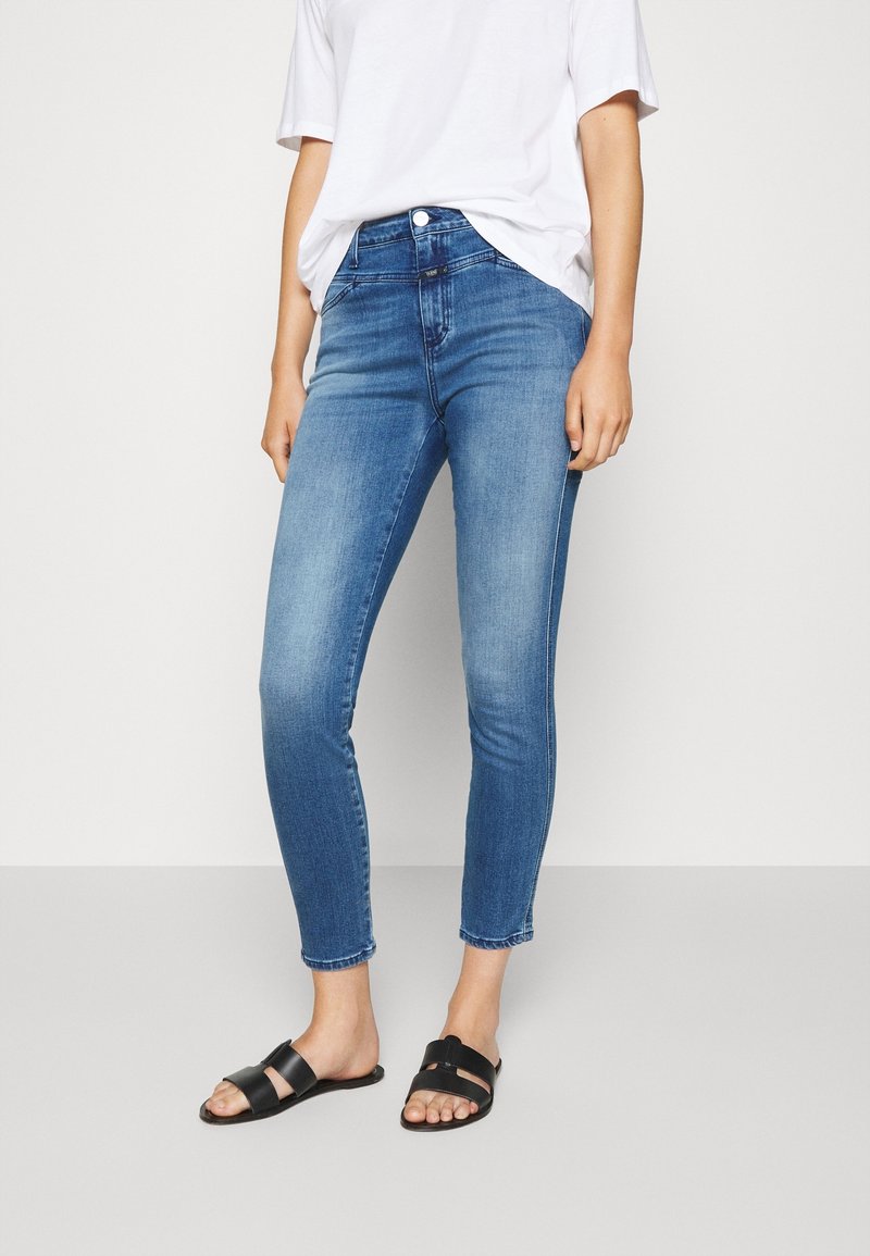 Jean en denim bleu clair à coupe slim et longueur cheville, associé à un t-shirt blanc ample et des sandales noires à enfiler.