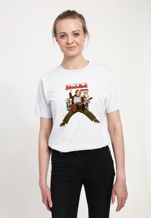Mladá žena v bílem tričku s potiskem "School of Rock" zobrazujícím postavu hrající na kytaru a skupinu dětí v pozadí, černé kalhoty.