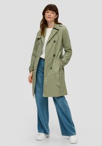 Jasnozielony płaszcz trench z podwójnym zapięciem, dużymi guzikami i bocznymi kieszeniami. W zestawieniu z szerokimi niebieskimi dżinsami i białymi butami.
