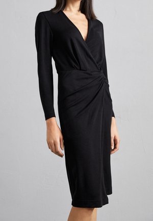 Kobieta ubrana w czarną, długorękawową sukienkę do kolan z dekoltem w kształcie litery V i marszczeniem w talii, na szarym tle.