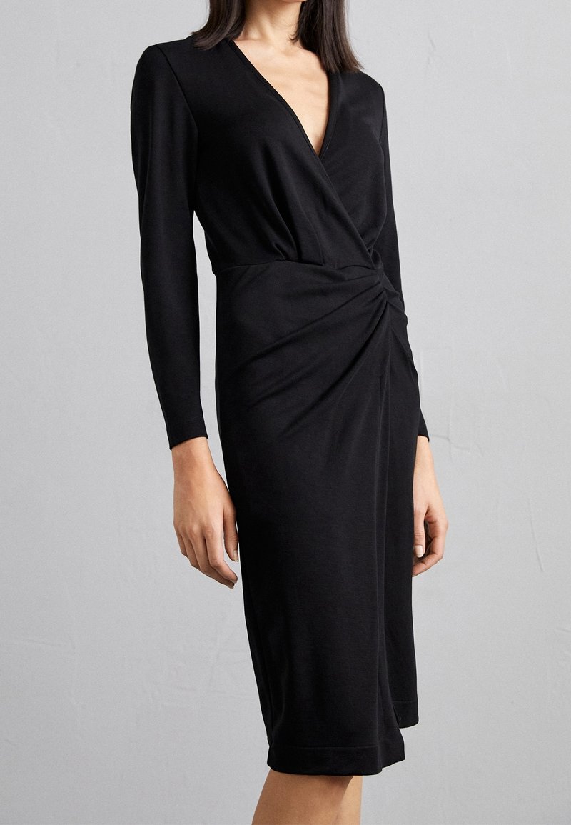 Femme portant une robe noire à manches longues, longueur genou, avec un décolleté en V et un détail froncé à la taille, sur un fond gris.