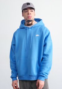 Helle blaue Baumwollkapuzenjacke mit einer Fronttasche, Kordeln und einem weißen Nike-Logo auf der Brust. Getragen mit grauen Hosen.