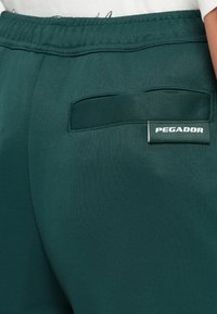 Zöld atlétikai rövidnadrág, textúrált, rugalmas anyagból készült. Jellemzője egy gumírozott derék és egy hátsó zseb, amelyen egy fehér "PEGADOR" felirat található.
