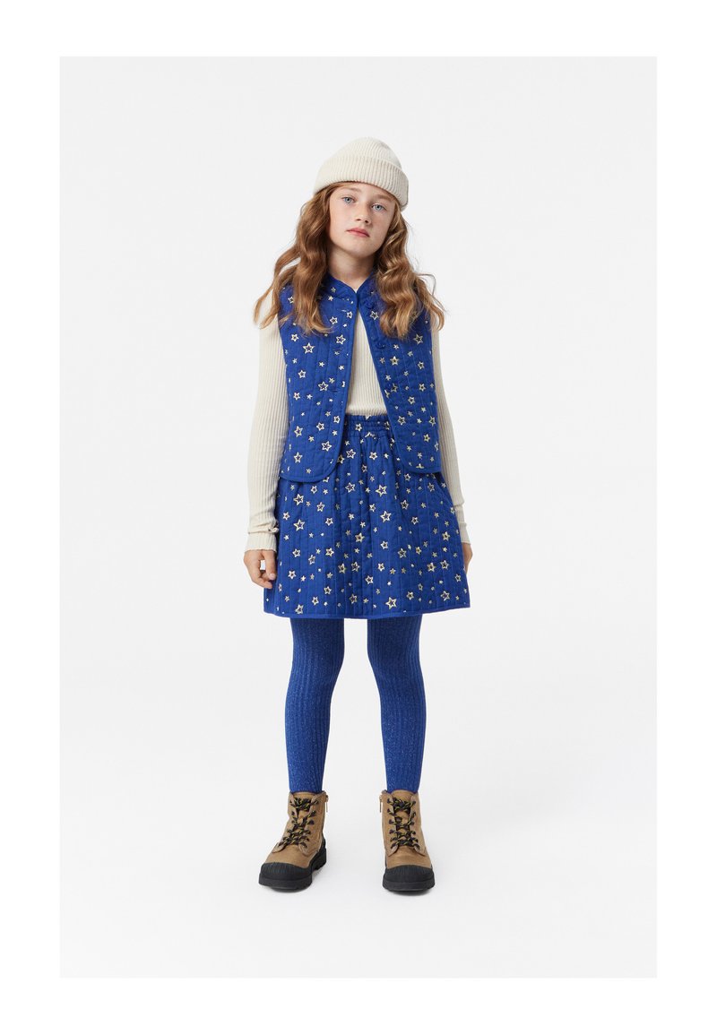 Blå vadderad väst med stjärnmönster, matchande kjol, ribbade blå leggings och bruna snörskor. Modellen har på sig en ljus stickad mössa.