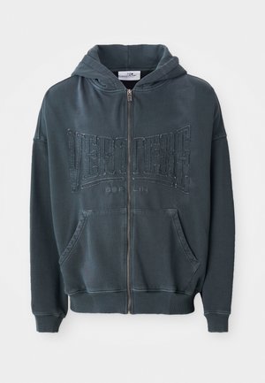 Haut de hoodie zippé vert foncé avec un logo texturé "VERTETRE BERLIN" sur le devant, doté d'une capuche avec cordon de serrage et de poches kangourou.