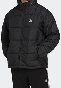 Svart puffjacka med hög krage, dragkedja och vadderad design. Har Adidas-logotypen i vitt på vänstra bröstet.