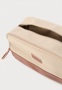 Μπεζ υφασμάτινη τσαντούλα καλλυντικών με καφέ δερμάτινη βάση και φερμουάρ, μερικώς ανοιχτή, που δείχνει κενό εσωτερικό σε λευκό φόντο.