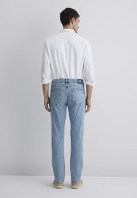 Light blue jeans with a straight-leg design, featuring a back patch pocket and a logo tag on the waistband, paired with a white shirt.
