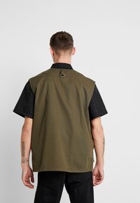 Khakiveste mit geradem Schnitt, mit Kragen und einer Aufhängeschlaufe hinten, getragen über einem kurzärmeligen karierten Hemd.