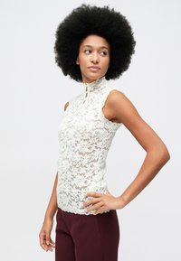 Kvinne med stor afrofrisyre, iført ermeløs hvit blomstermønstret bluse i blonde og maroon bukser, ser sidelengs med hånden på hoften.