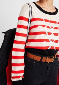 Červeno-krémový pruhovaný svetr s černým výstřihem, zdobený srdíčkem. Spárován s tmavými džínami a hnědým páskem. Černá bunda leží poblíž.