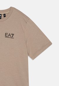 Bežová bavlnená tričko s okrúhlym výstrihom a krátkymi rukávmi, s čiernym logom EA7 Emporio Armani na prednej strane. Hladká textura.