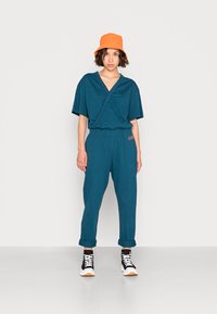 Teal wikkeltop met korte mouwen en bijpassende taps toelopende broek, met een elastische tailleband. Draagt een oranje bucket hat en zwarte sneakers.