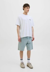 Hvid bomulds t-shirt med kortærmer og et lille sort logo, parret med lyseblå denim shorts, hvide ankelstrømper og beige sko.