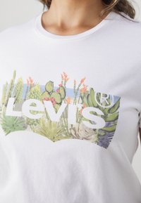 T-shirt en coton blanc avec un grand graphique coloré de cactus et de fleurs, affichant en gros caractères blancs le logo de la marque "Levi's".
