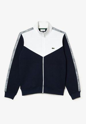 Veste zippée bleu marine et blanche avec col montant, logo Lacoste sur la poitrine et bandes marquées sur les manches.