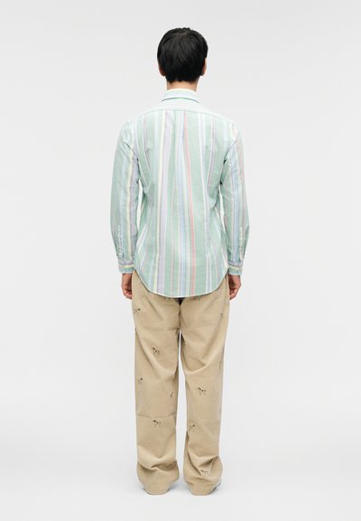 Οπίσθια όψη ενός άνδρα που φοράει ανοιχτοπράσινο πουκάμισο με ρίγες και μπεζ παντελόνι με κεντητά σχέδια. Το πουκάμισο έχει μακριά μανίκια και καμπυλωτό τελείωμα.