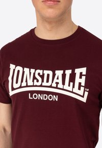 Burgundi pamut póló, amelyen hangsúlyos fehér "Lonsdale London" logó található, vastag, nagybetűs betűtípussal, klasszikus kerek nyakkivágással.