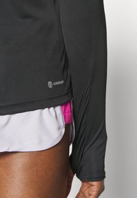 Musta pitkähihaiseksi urheilu paita ribbitekstuurilla, jonka helman lähellä on pieni valkoinen Aeroready-logo. Kirkkaan vaaleanpunaiset shortsit näkyvät osittain alla.