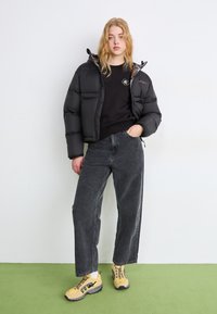 Giacca a vento nera con zip e cappuccio, abbinata a una felpa nera e jeans grigi a vita alta. Presenta sneaker gialle con dettagli neri.