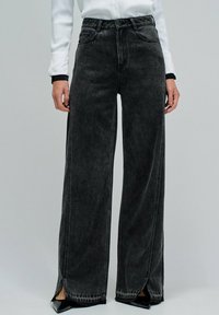 Szersze czarne dżinsy z wyblakłym wykończeniem, z kieszeniami z przodu i rozcięciami po bokach na dole. Zaprojektowane w standardowym stylu pięciu kieszeni.