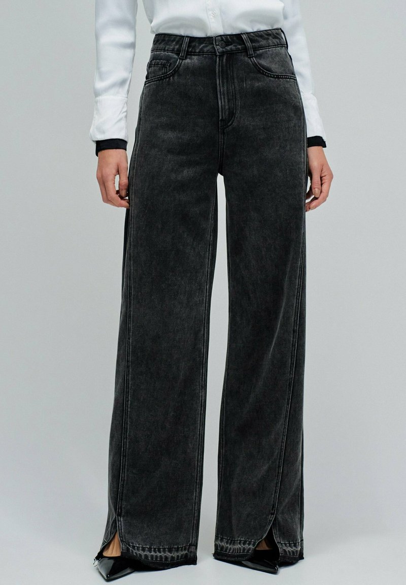 Szersze czarne dżinsy z wyblakłym wykończeniem, z kieszeniami z przodu i rozcięciami po bokach na dole. Zaprojektowane w standardowym stylu pięciu kieszeni.