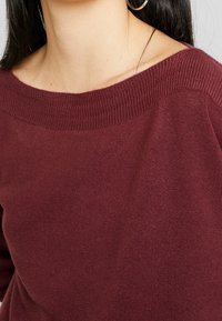 Maglione bordeaux a maglia con un ampio scollo a coste e una texture morbida. Dettaglio in primo piano che mette in risalto la fine tessitura del materiale e il colore.
