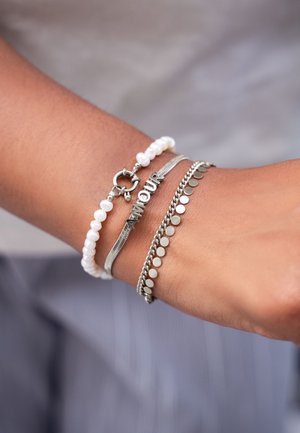 Muñeca con tres pulseras: una de cuentas blancas, una cadena plateada con las letras "AMOUR" y una cadena plateada con pequeños discos redondos.