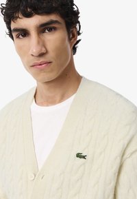 Cardigan color crema con texture a maglia a treccia, scollo a V profondo e chiusura con bottoni, caratterizzato da un piccolo logo di coccodrillo verde sul lato sinistro del petto.