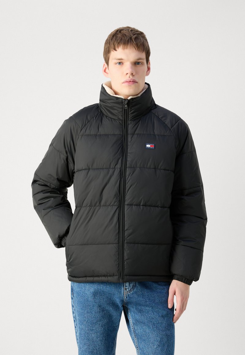 Schwarze Pufferjacke mit hohem Kragen, gesteppter Optik und Reißverschluss. Verfügt über ein LOGO-Patch und elastische Bündchen. Getragen über blauen Denim-Jeans.