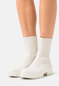 Botas de tobillo de cuero blanco con una textura suave, puntera redondeada y suela de goma maciza con profundos surcos. Diseño sin costuras y acentos mínimos.