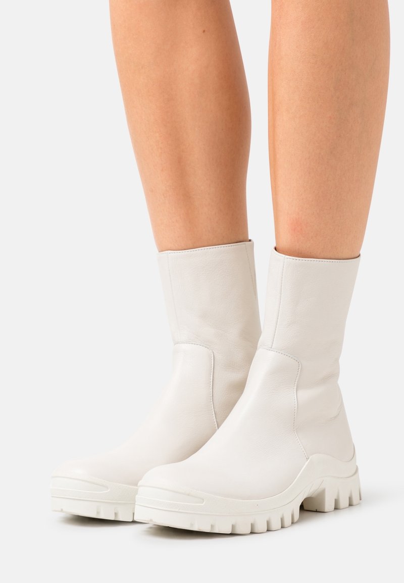 Botas de tobillo de cuero blanco con una textura suave, puntera redondeada y suela de goma maciza con profundos surcos. Diseño sin costuras y acentos mínimos.
