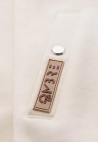 Tissu crème avec une texture lisse, présentant une étiquette rectangulaire marron avec un texte en relief et un détail de bouton argenté.