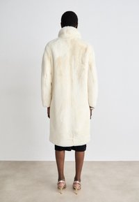 Lang, cremet faux fur frakke med høj krave, der har en blød tekstur og et minimalistisk design, og som slutter midt på læggen. Inkluderer en lige pasform og sidelommer.