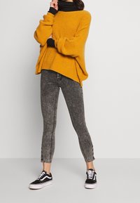 Maglione a maglia oversize giallo senape con scollatura ampia, abbinato a jeans skinny grigi a vita alta e sneakers bianche e nere.