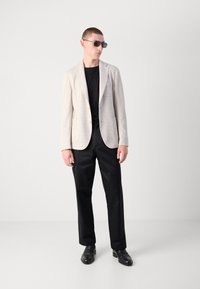 Blazer beige con texture sopra una maglietta nera, abbinato a pantaloni neri e scarpe nere. Accessoriato con occhiali da sole scuri.