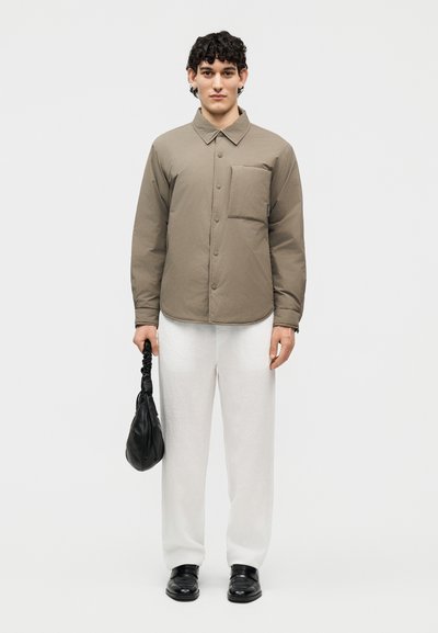 Young man wearing a taupe buttoned shirt with chest pocket, white pants, black shoes, and holding a small black handbag against a plain background.