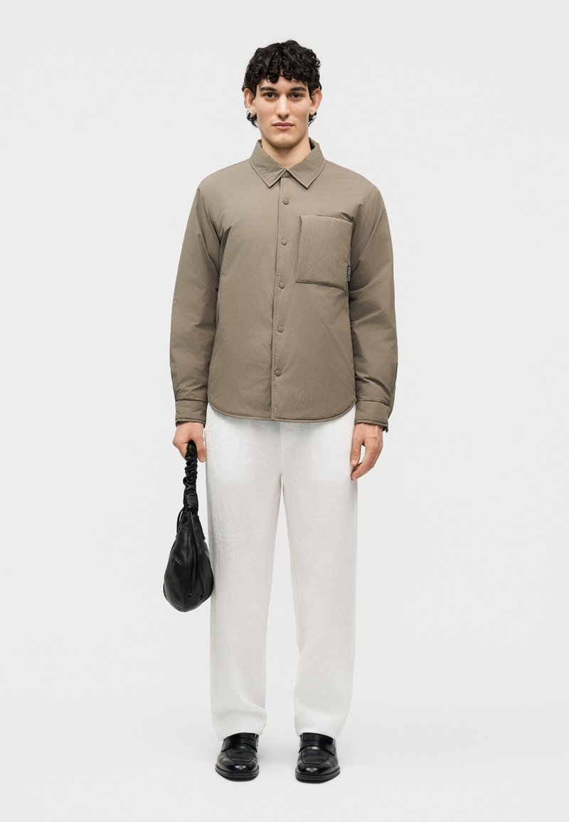 Jeune homme portant une chemise boutonnée taupe avec poche poitrine, pantalon blanc, chaussures noires, et tenant un petit sac à main noir devant un fond uni.