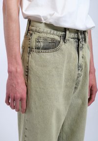 Bleka gröna denimjeans med hög midja, två framfickor och en avslappnad passform. Synlig sömnad och lätt slitning vid kanterna.