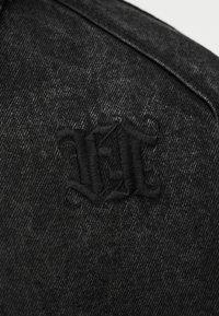 Μαύρο τζιν ύφασμα με μαύρα κεντημένα αρχικά "M" σε γοτθική γραμματοσειρά κοντά σε μια ραφή.