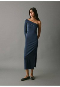 Vestito blu navy scollato sulle spalle con uno spacco laterale, caratterizzato da accenti circolari decorativi lungo lo scollo. Tessuto morbido ed elastico. Scarpe basse nere.