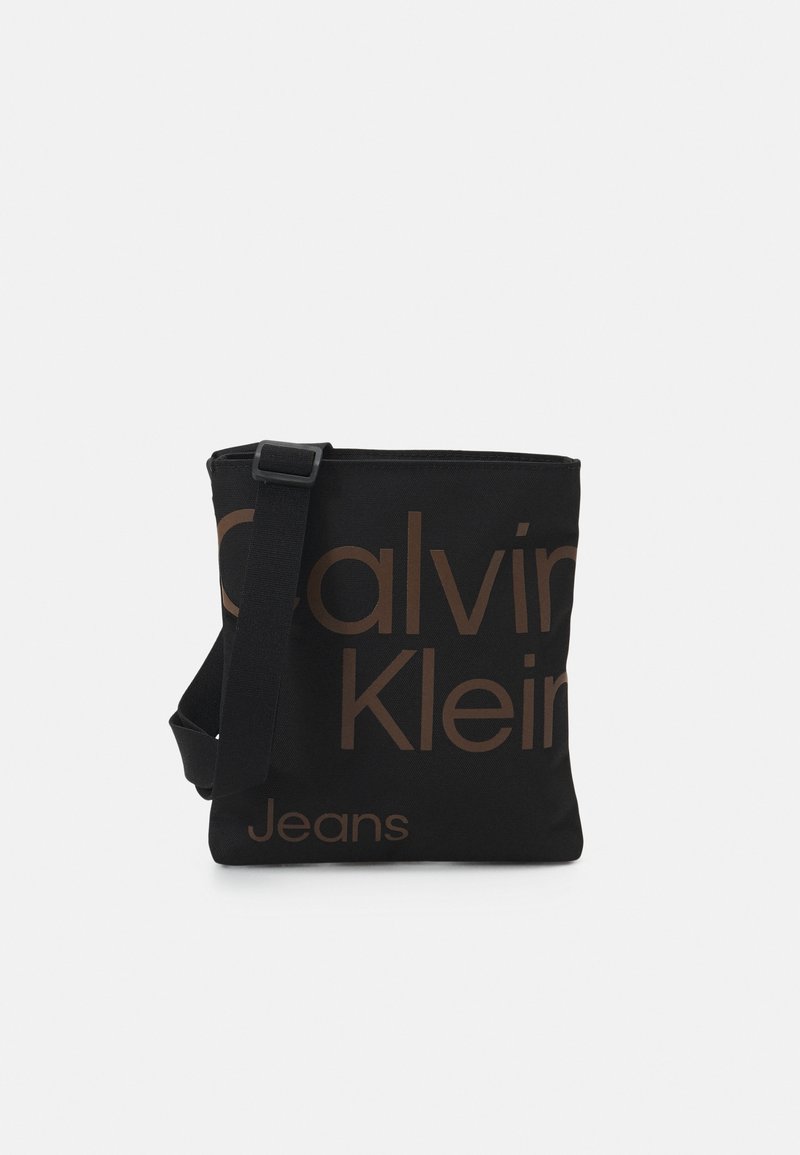 Svart canvas crossbody-väska med fet brun text "Calvin Klein Jeans". Har en justerbar rem och en dragkedja upptill.