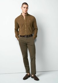 Bruin overhemd met knopen, textuur bruin geruit broek en bruine schoenen maken een smart-casual outfit compleet. Model poseert met één hand in de zak.