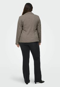 Žena s dlouhými hnědými vlasy, oblečená v hnědém kostkovaném saku a černých bootcut džínách, stojí zády k fotoaparátu na bílem pozadí.