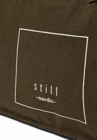 Canvas-Tragetasche in dunkelbraun mit einem weißen quadratischen Rand und dem Text "still nordic." Glatte Textur mit verstärkten Nähten.